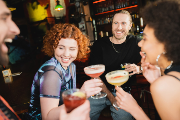 Positieve man met blauwe lagune cocktail in de buurt van interraciale vrienden praten in de bar  - Foto, afbeelding