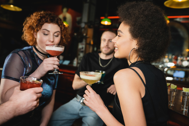 Positive curly african american woman holding pisco sour cocktail near friends in bar  - Photo, Image