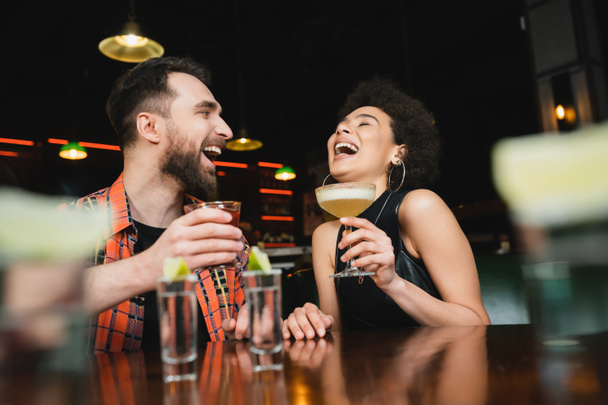 Laughing african american woman holding cocktail near friend and tequila shots in bar  - Foto, Imagem