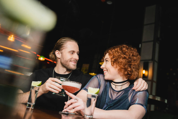 Positive man hugging red haired friend with cocktail near stand in bar  - 写真・画像