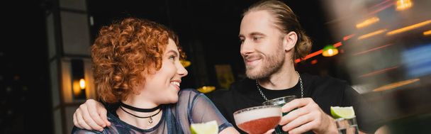 Smiling bearded man hugging redhead friend with cocktail in bar, banner  - Photo, Image