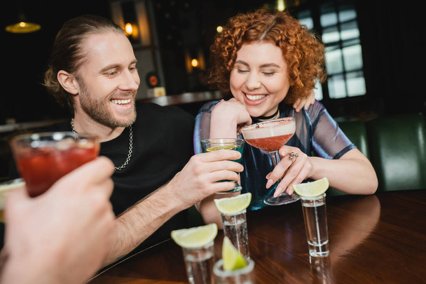 Χαμογελώντας φίλους clinking διαφορετικά κοκτέιλ κοντά σφηνάκια τεκίλα με ασβέστη στο μπαρ  - Φωτογραφία, εικόνα
