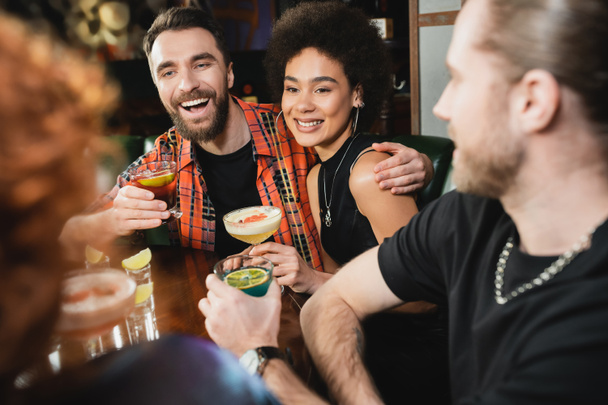 Positive multiethnic people holding different cocktails near blurred friends in bar  - Photo, Image