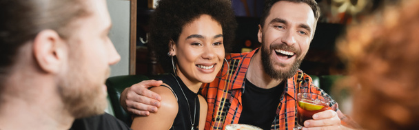 Un homme souriant tenant un cocktail negroni et embrassant un ami afro-américain dans un bar le soir, bannière  - Photo, image