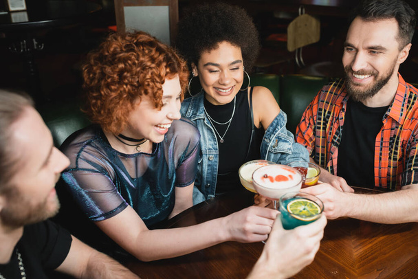 Smiling interracial women holding cocktails near bearded friends in bar  - Fotografie, Obrázek