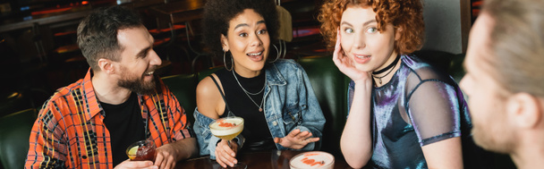 Emocionadas mujeres interracial sosteniendo cócteles cerca de hombres borrosos en el bar, pancarta  - Foto, imagen