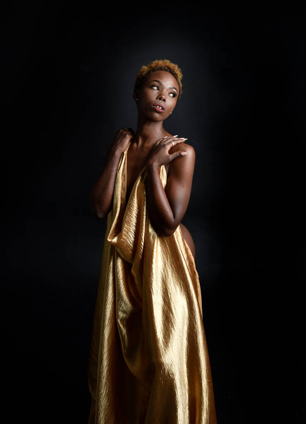  アフロ、女神シルクのローブと華やかな花冠と美しいアフリカの女性モデルの幻想的な肖像画。ジェスチャー黄金の花を保持ポーズ。暗いスタジオの背景に隔離され  - 写真・画像