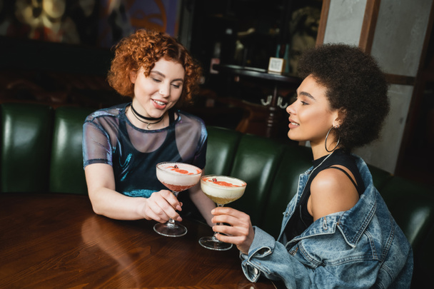 Redhead woman holding clover club cocktail and talking to african american friend in bar   - Fotografie, Obrázek