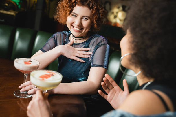 Sorglose Frau hält Klee-Club-Cocktail neben verschwommener afrikanisch-amerikanischer Freundin in Bar  - Foto, Bild