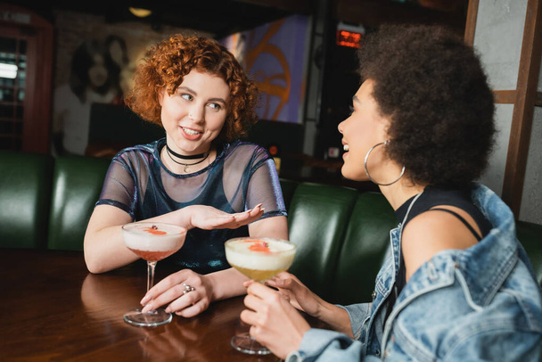 Uśmiechnięta rudowłosa kobieta rozmawia z afrykańską dziewczyną koło koktajli w barze  - Zdjęcie, obraz