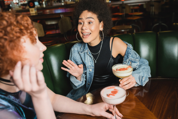 Femme afro-américaine excitée parlant à un ami flou près de cocktails en mousse dans le bar  - Photo, image