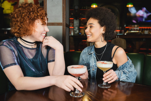 Jóvenes novias multiétnicas hablando cerca de cócteles de espuma en el bar  - Foto, Imagen