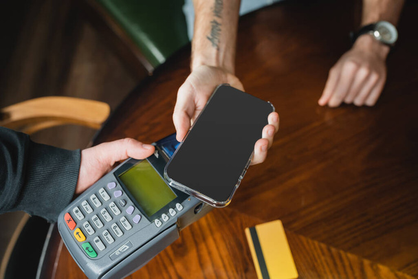 Ausgeschnittene Ansicht von Mann, der mit Smartphone in der Nähe von Kellner mit Zahlungsterminal in Bar bezahlt  - Foto, Bild