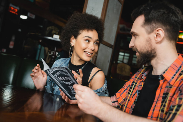 Sorrindo afro-americana falando com amigo barbudo com menu no bar  - Foto, Imagem