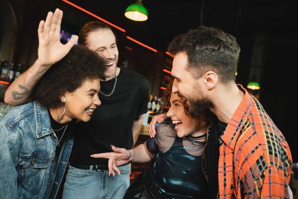 Excited man waving hand near interracial friends while spending time in bar at night  - Photo, Image