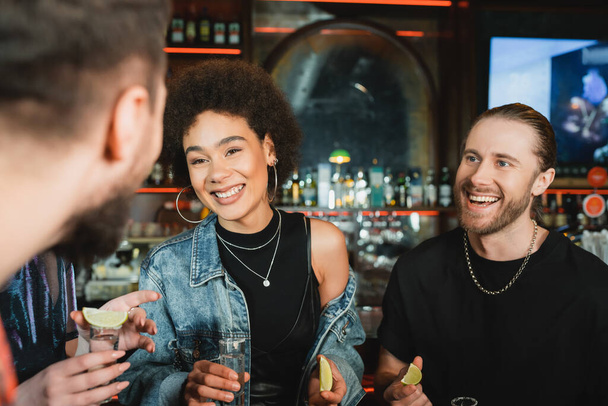 Fröhliche afrikanisch-amerikanische Frau hält Limetten und Tequila neben Freunden in Bar  - Foto, Bild