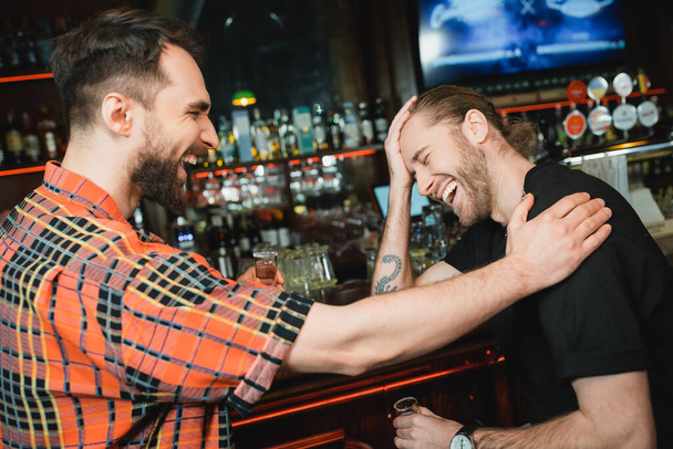 Cheerful bearded men holding tequila shots while laughing in bar in evening  - Photo, Image