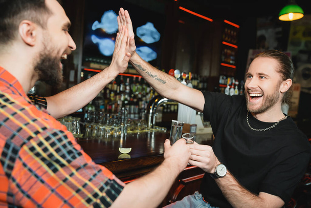 Positive bearded friends giving high five and clinking tequila in bar  - Photo, Image
