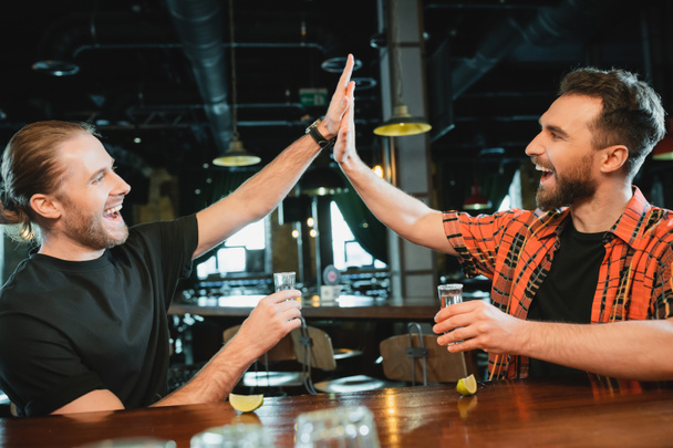 Ενθουσιασμένοι φίλοι να δίνουν κόλλα πέντε και να κρατούν σφηνάκια τεκίλα κοντά σε ώριμο λάιμ στο μπαρ  - Φωτογραφία, εικόνα