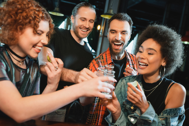 Carefree multiethnic friends holding lime and clinking tequila with salt in bar  - Foto, Imagem