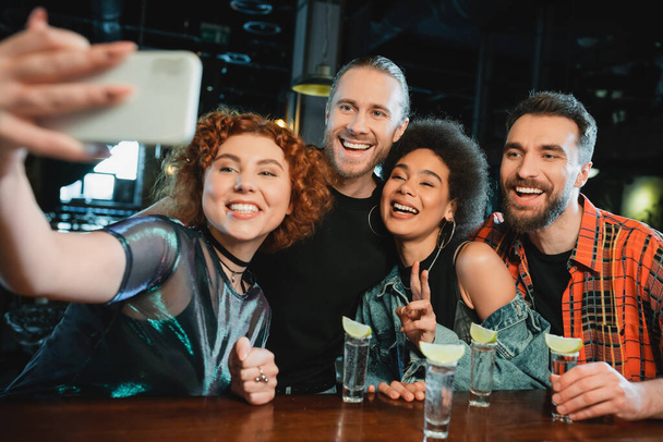 Smiling multiethnic friends taking selfie on smartphone near tequila in bar  - Photo, Image