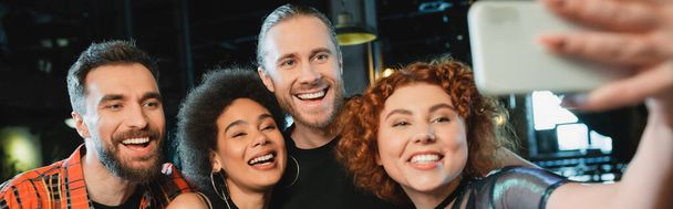 Young redhead woman taking selfie with interracial friends in bar, banner  - Photo, image