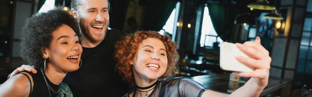 Overjoyed redhead woman taking selfie on smartphone near multiethnic friends in bar, banner  - Фото, изображение