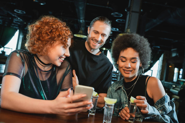 Usmívající se zrzka žena drží smartphone v blízkosti multietnických přátel a tequila v baru  - Fotografie, Obrázek