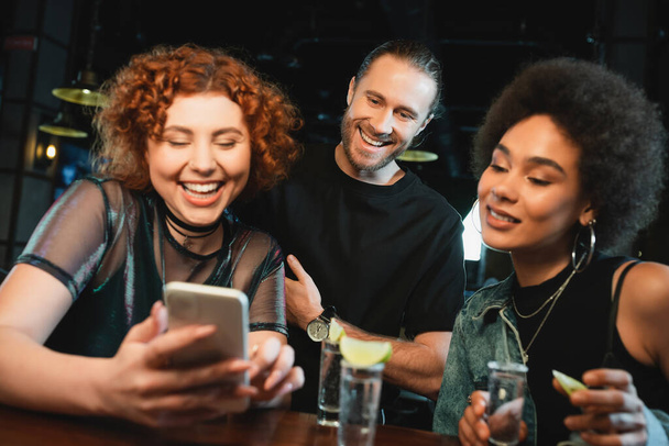 Cheerful multiethnic friends using smartphone near tequila shots in bar  - Foto, Bild
