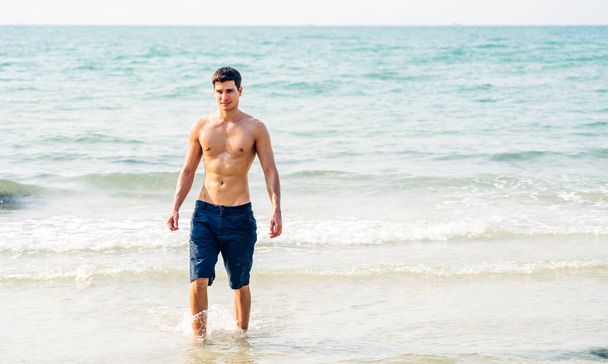 Gülümseyen yakışıklı seksi adamın portresi kaslı fit vücudunu gösteriyor tropik sahilde durup eğleniyor ve rahatlıyor. - Fotoğraf, Görsel