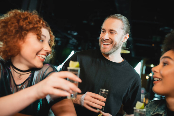Sonriente hombre barbudo mirando borrosa interracial amigos con tequila en el bar  - Foto, imagen