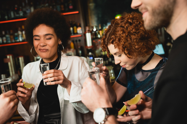 african american woman grimacing after tasting sour lime near multiethnic friends holding tequila shots in bar  - 写真・画像