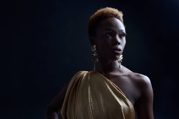  fantasy portret pięknej afrykańskiej modelki z afro, boginią jedwabnych szat i ozdobną wieńcową koroną. gestu pozowanie trzymając złote kwiaty. izolowany na ciemnym tle studio  - Zdjęcie, obraz