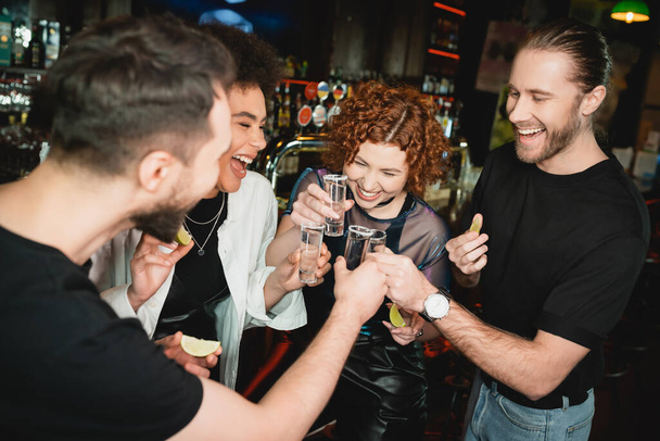 Smiling redhead woman clinking tequila shots with interracial friends in bar  - Foto, afbeelding