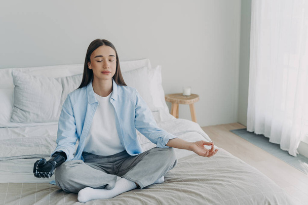 Meditation and concentration. Morning routine of disabled woman. Girl is sitting in lotus pose in her bedroom. Attractive european woman has artificial cyber limb. Stress relief at home. - Foto, Bild