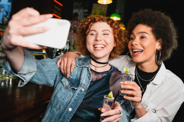 Mujer positiva tomando selfie con amigo afroamericano y sosteniendo tequila en el bar  - Foto, imagen
