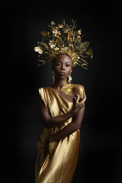  πορτρέτο φαντασίας του όμορφου αφρικανικού γυναικείου μοντέλου με αφρό, μεταξωτές ρόμπες θεάς και περίτεχνο στεφάνι στεφανιού. χειρονομιακή ποζάρισμα εκμετάλλευση χρυσά λουλούδια. απομονωμένο σε σκοτεινό φόντο στούντιο  - Φωτογραφία, εικόνα