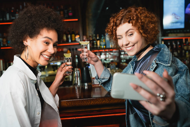 Pozytywne wielonarodowe dziewczyny trzymające tequilę z limonką i robiące selfie w barze  - Zdjęcie, obraz