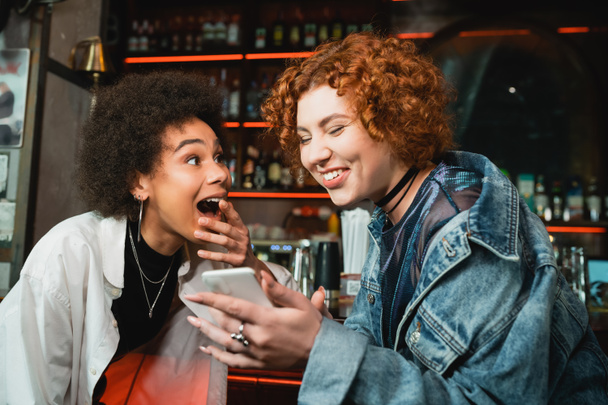 Excited redhead woman using smartphone near african american friend in bar  - Fotografie, Obrázek