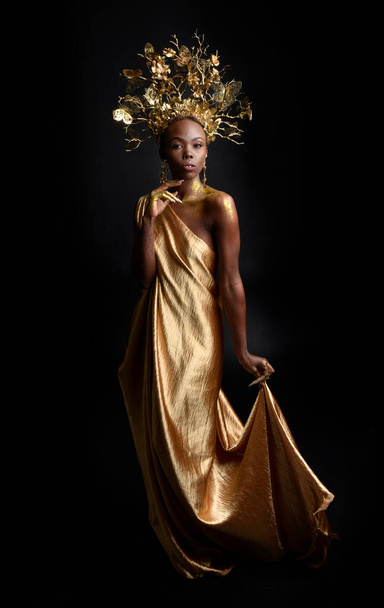  Afro, tanrıça ipek cübbesi ve süslü çiçek tacıyla güzel Afrikalı kadın modelinin fantezi portresi. Altın çiçekleri tutan hareketli pozlar. Karanlık stüdyo arka planında izole  - Fotoğraf, Görsel