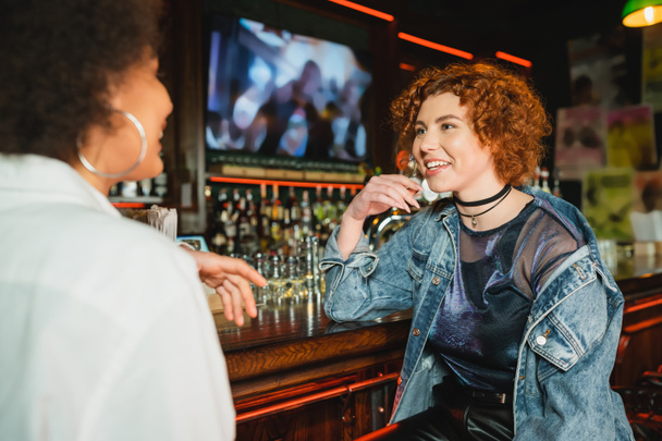 Mulher ruiva jovem falando com amigo americano africano borrado perto de stand no bar  - Foto, Imagem