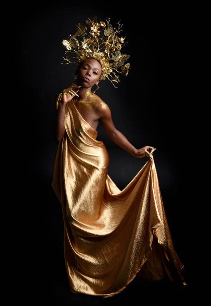  fantasy portrait of beautiful african woman model with afro, goddess silk robes and ornate floral wreath crown. gestural Posing holding golden flowers. isolated on dark  studio background  - Photo, Image