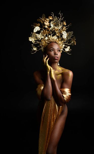  Afro, tanrıça ipek cübbesi ve süslü çiçek tacıyla güzel Afrikalı kadın modelinin fantezi portresi. Altın çiçekleri tutan hareketli pozlar. Karanlık stüdyo arka planında izole  - Fotoğraf, Görsel