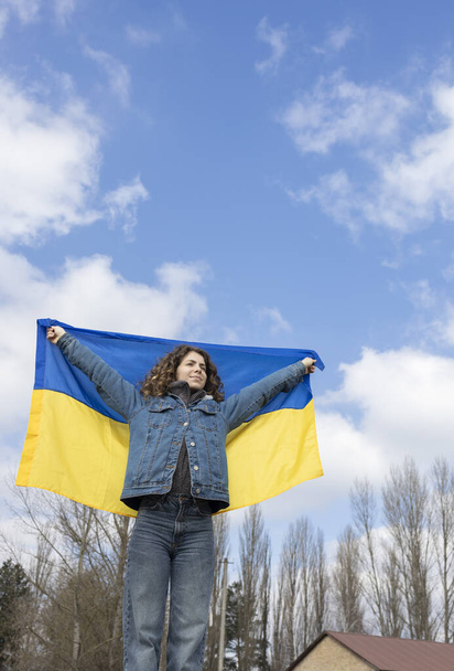 fiatal nő, egy felnőtt lány, egy ukrán sárga-kék zászlót tart a háta mögött az égnek. Az ukránok ellenzik a háborút. Felhívja a figyelmet az ukrajnai katonai műveletekre. Állj Ukrajna mellé! - Fotó, kép