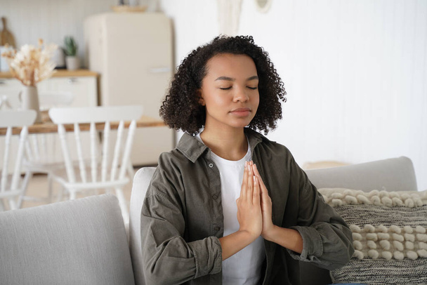 Teenage african american girl is practicing yoga at home. Curly young spanish woman is sitting on couch in lotus pose with her eyes closed. Harmony and balance. Wellness and spirituality. - Valokuva, kuva