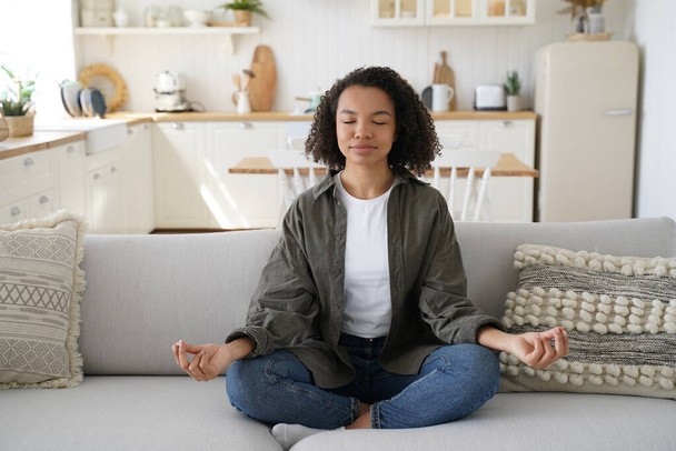 Teenage afro girl is practicing yoga at cosy home. Young woman sitting on couch in lotus pose. Concentration and balance. Wellness and emotional wellbeing, tranquility and recreation, stress relief. - Foto, immagini