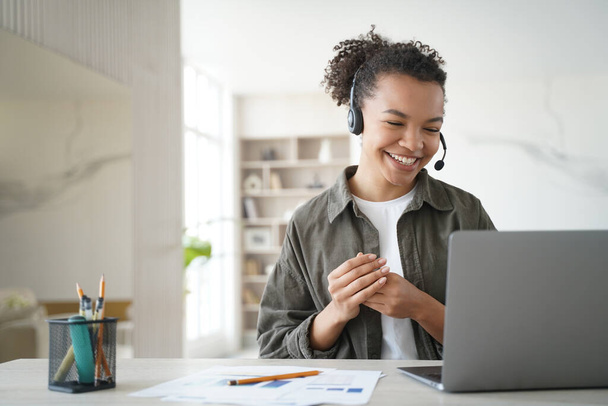 Happy diák online leckét kap otthonról. Távolsági tanulmány a karanténról. Egy fiatal nő fülhallgatóban tanácskozik. Az afro-amerikai menedzser mosolyog. Támogatási szolgáltatás üzemeltetőjének távolról végzett munkája. - Fotó, kép