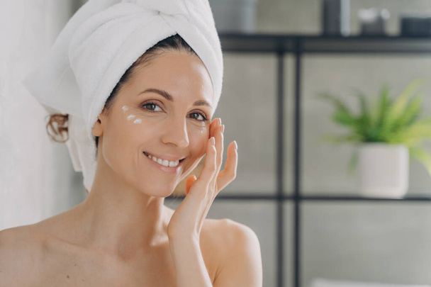 Face of beautiful european lady which applies cream. Attractive feminine caucasian girl wrapped in towel after bathing and hair washing. Happy young woman takes shower at home. Beauty routine. - Valokuva, kuva