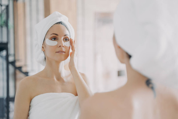 Mirror reflection of girl which applies anti wrinkle eye patches and doing skincare. Attractive caucasian woman wrapped in towel after bathing and hair washing. Relaxation at spa resort or at home. - Photo, Image