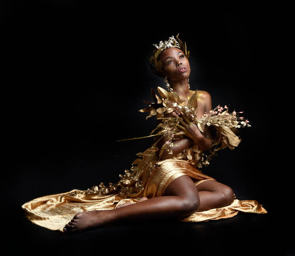  fantasy portret pięknej afrykańskiej modelki z afro, boginią jedwabnych szat i ozdobną wieńcową koroną. gestu pozowanie trzymając złote kwiaty. izolowany na ciemnym tle studio  - Zdjęcie, obraz
